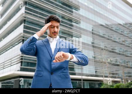 A Hispanic businessman in a blue suit looks concerned while checking the time on his watch outside a corporate building, representing the fast-paced nature of business, feels leak of time and deadline Stock Photo