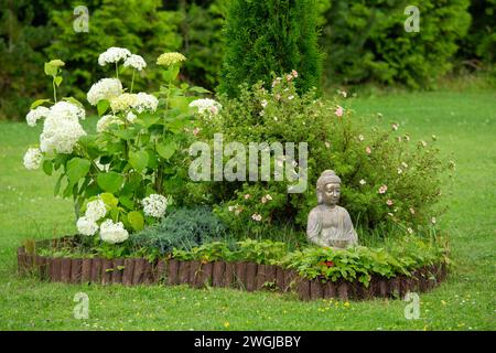 Meditating Buddha figure sit inside small island of flowers, Potentilla bush and hydrangea in flower bed, surrounded by natural wood roll palisade. Stock Photo