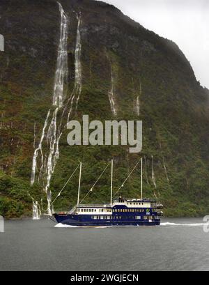 Waterfalls and the Fiordland Navigator cruise ship making way on a rainy day in Doubtful Sound / Patea, Fiordland /Te Rua-o-te-Moko, New Zealand / Aot Stock Photo