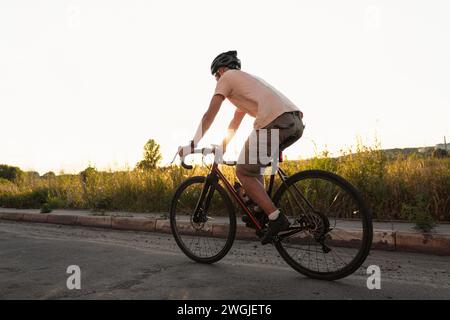 Young sporty guy in a helmet rides on gravel bicycle on asphalt road at sunset. Outdoor cycling training on fresh air. Stock Photo