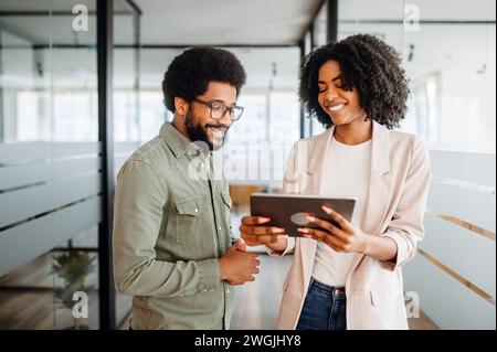 Two cheerful colleagues are sharing a tablet screen, the woman pointing at the screen, a man watching with interest, collaborative and friendly work atmosphere. Team interaction and knowledge exchange Stock Photo