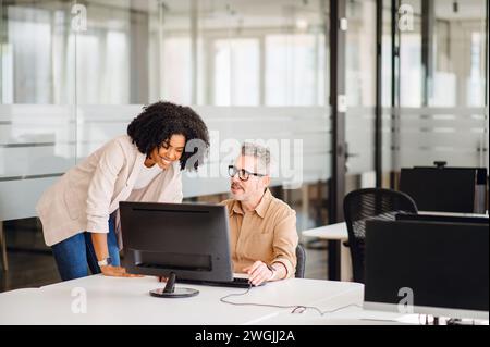 Cheerful woman in business casual attire leaning over a male colleague, sharing ideas or advice. Their easy rapport suggests a brainstorming session in a supportive and collaborative office atmosphere Stock Photo