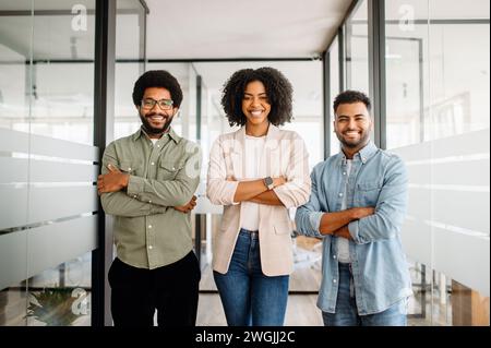 Three vibrant colleagues stand together in an office, their arms crossed, signifying unity and a strong team dynamic. The diverse and cohesive group, ready for collaborative success. Stock Photo