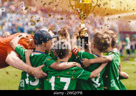 Happy children in sports team celebrating victory. Kids winning the game and rising golden cup. Kids football team huddling in circle and jumping in h Stock Photo