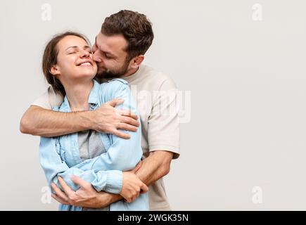 Happy young woman in the arms of her husband in front of light grey background win studio. Man hugs woman. Stock Photo