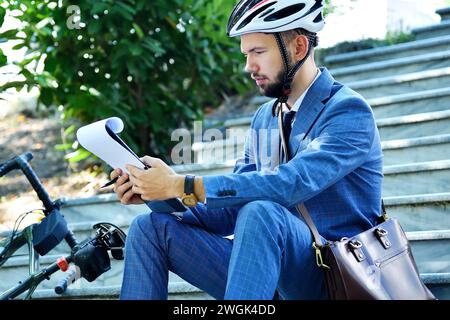 Young businessman in helmet sitting on stairs and analyzes business report in clipboard. Business and working concept. Stock Photo