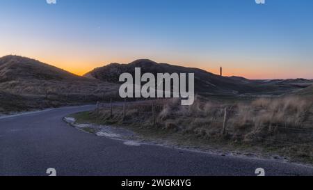 Asphalt road through the dunes of the Netherlands during sunrise. On the horizon, not only is the lighthouse visible, also the crescent moon and Venus Stock Photo