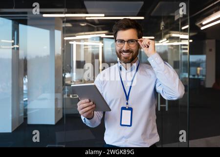 Portrait of mature experienced programmer system administrator, man smiling and looking at camera with tablet computer in hands, mature worker testing new software inside office at workplace. Stock Photo