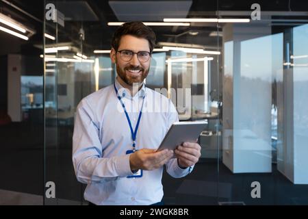 Portrait of mature experienced programmer system administrator, man smiling and looking at camera with tablet computer in hands, mature worker testing new software inside office at workplace. Stock Photo