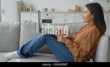 Side view Asian woman lying on couch at home korean japanese businesswoman girl typing on laptop chatting online search data read social media Stock Photo