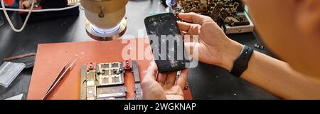 cropped view of skilled repairman holding mobile phone with broken screen near devices, banner Stock Photo