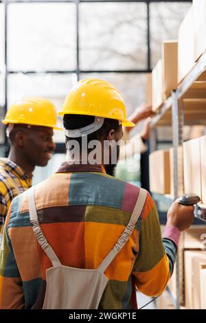 Merchandise managers analyzing stock inventory using barcode scanner in warehouse. African american storehouse employees wearing protective helmets and overalls scanning cargo qr code Stock Photo