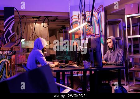 Internet criminals working together to break into database in dark room. African american and asian young men hacking computer system and installing virus malicious software Stock Photo