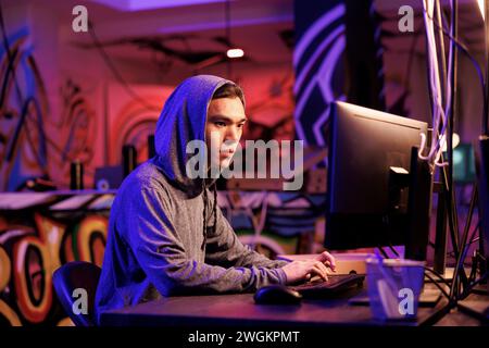Asian internet scammer cracking password on computer to get access to database. Young man in hood writing malicious software code on computer and breaking into server in dark room Stock Photo