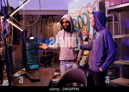 Hackers discussing hacking plan and cybercrime technique. Criminals in hood standing near computer and talking about malicious software code on screen in abandoned warehouse Stock Photo