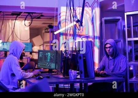 Hackers collaborating while coding virus software, doing illegal activities and breaking law in abandoned warehouse. Criminals programming malware on computers in hideout place Stock Photo