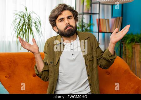 I dont know what to say. Confused middle eastern Arabian man feeling embarrassed about ambiguous question, having doubts, no idea, being clueless and uncertain. Young bearded guy at home room on couch Stock Photo