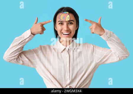 Beautiful young happy woman pointing at paper fish attached to her forehead on blue background. April Fools Day celebration Stock Photo