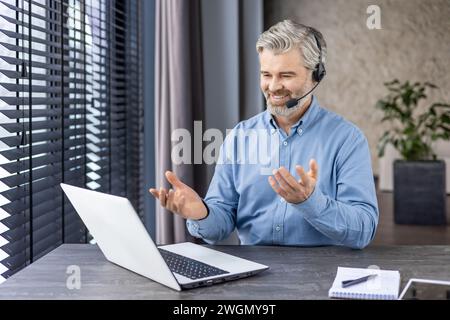 Elderly grey-haired man in denim shirt sitting at domestic workspace with laptop and having distant meeting with coworkers. Caucasian businessman in headset using online communication privileges. Stock Photo