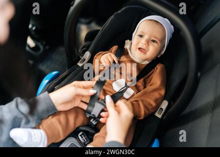 Close-up high-angle view of unrecognizable young mother fastening safety belt to unhappy infant baby boy in car seat. Crying toddler with security Stock Photo
