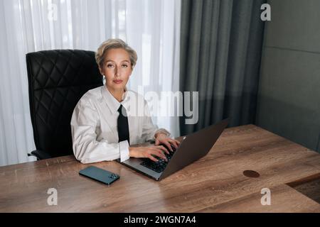 High-angle view portrait of serious focused mature 50s businesswoman looking at camera, typing on laptop keyboard, working on online project Stock Photo