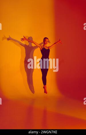 Athletic Grace. Body-Size Portrait of beautiful woman, ballerina dancer stretches in black sports overalls, barefoot in neon-lit studio. Stock Photo