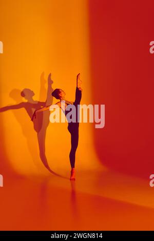 Flexibility in Neon Glow. Beautiful woman stretches gracefully in black sports overalls, barefoot in neon-lit studio, shadow mirroring her movements. Stock Photo