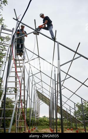 Man in safety helmet standing on ladder and holding hammer while colleague installing metal rail for solar modules. Male workers assembling support structures for solar panels under white sky. Stock Photo