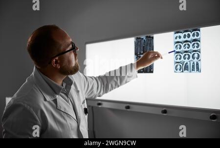 Side view of male doctor studying MRI results. Male specialist working in hospital, carrying out description of the radiographs of patient, pointing by pen. Specialist wearing glasses and white robe. Stock Photo
