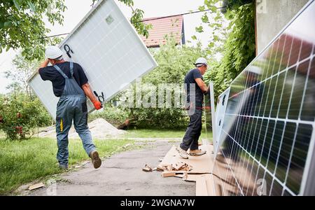 Two men carrying solar modules in the yard. Male worker in safety helmet holding photovoltaic solar panel and walking on grass while colleague standing by house with solar module. Stock Photo