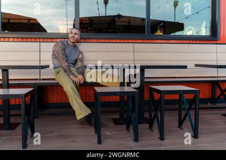 A handsome young man with tattoos, sitting on a bench at an outdoor restaurant Stock Photo