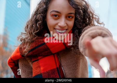 young mixed race smiling woman in coat and orange scarf  pointing her finger at the camera as in a screen Stock Photo