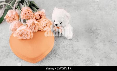 Heart-shaped peach fuzz velvet gift box with beautiful tulips and a teddy bear on a gray background, flat layer. Copy space. Stock Photo