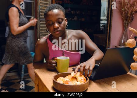 young afro woman at home studying with laptop taking croissant from bowl on table to eat with coffee while resting from studying. copy space. Stock Photo