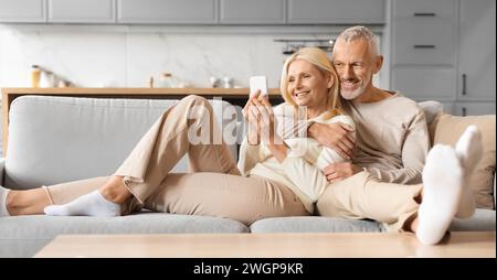 Relaxed senior spouses chilling on couch at home, using smartphone Stock Photo
