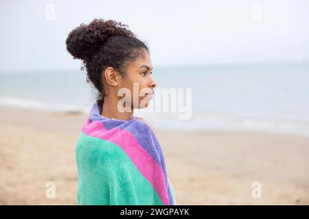 Portrait of a young mixed race woman on the beach, she is wrapped in a towel looking out to the ocean Stock Photo