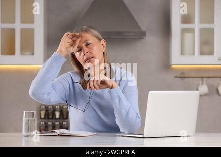 Menopause. Woman with glasses suffering from headache at table in kitchen Stock Photo