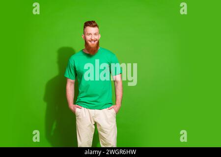 Photo of confident handsome man with ginger beard dressed green t-shirt holding hands in pockets isolated on green color background Stock Photo