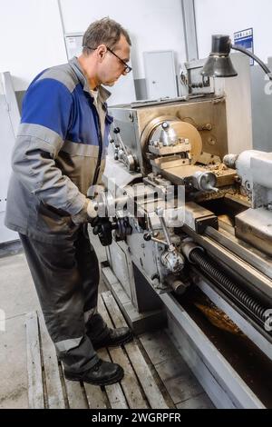 Turner in overalls works at lathe in turning shop at factory. Elderly worker turns metal part on machine. Side view Full height Stock Photo