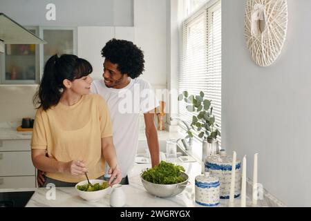 Happy young intercultural husband and wife in t-shirts looking at each other while woman mixing salad ingredients in bowl Stock Photo