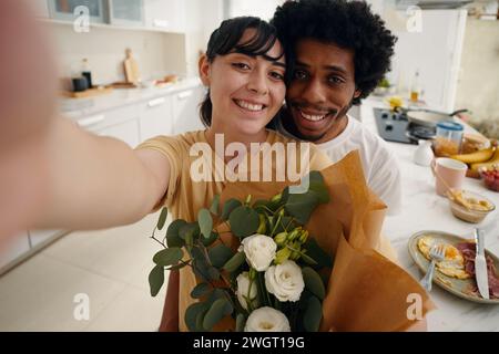 Young smiling man embracing his pretty wife with bunch of white flowers and both looking at camera while woman taking selfie Stock Photo
