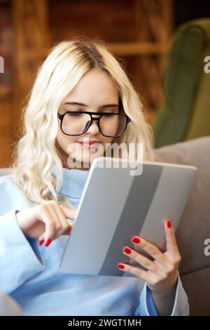 Portrait of young teen girl, student in protective glasses looking at tablet and studying online sitting on couch at home. Stock Photo