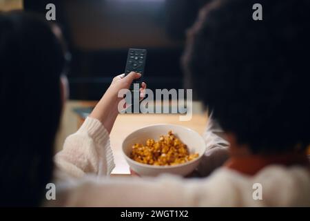 Hand of young woman pressing channel button on remote control while sitting next to her husband in front of TV set at leisure Stock Photo