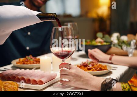 Young man pouring red wine into wineglass held by his wife sitting by served festive table in front of her husband in restaurant Stock Photo