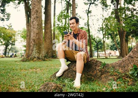 Young man with mobile phone and earphones sitting under big tree in park and scrolling through tracks in playlist at leisure Stock Photo