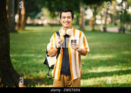 Young cheerful man in casualwear holding backpack on shoulder and smartphone in hand while standing in park with green lawns Stock Photo