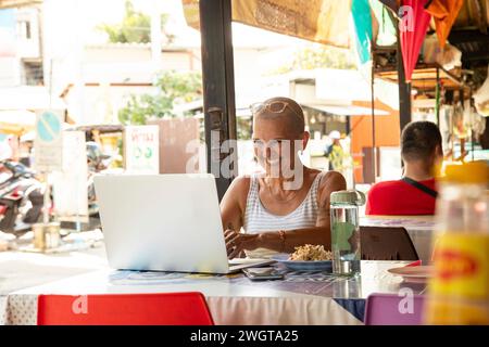 Mid aged woman solo traveller working on her laptop in a local restaurant in Chiang Mai. Thailand. she is smiling. Stock Photo