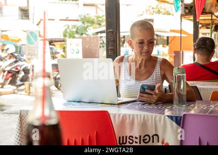 Mid aged woman solo travelle sits in a local restaurant using her phone and her laptop Stock Photo