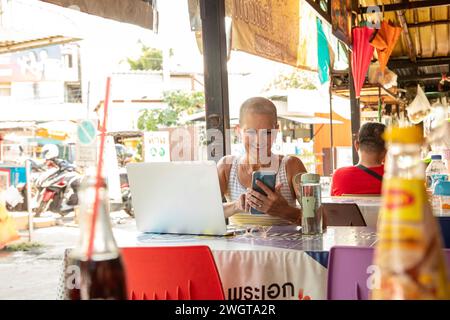 Mid aged woman solo travelle sits in a local restaurant using her phone and her laptop Stock Photo