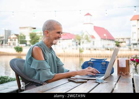 Mid aged woman sits in a riverside restaurant in Northern Thailand. She is on her laptop laughing. Stock Photo